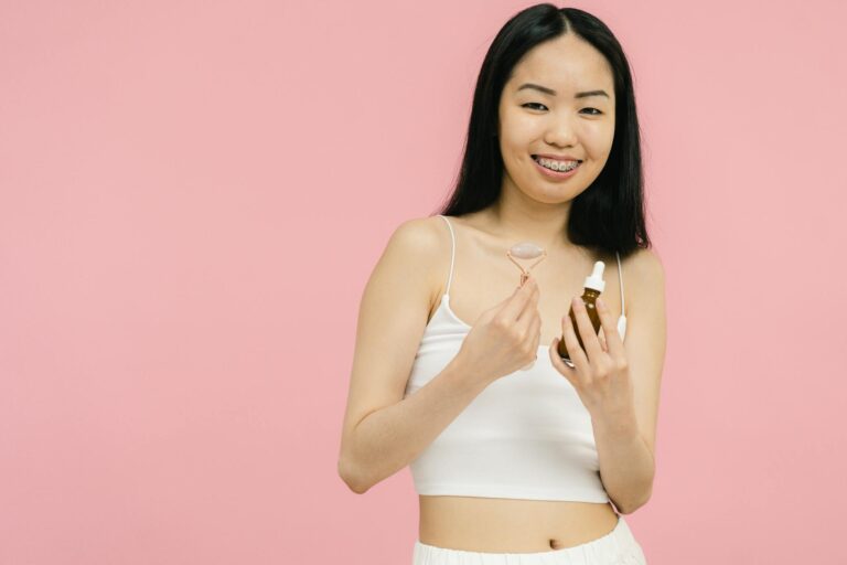 Woman smiling with facial roller and serum bottle against a pink backdrop, promoting skincare.