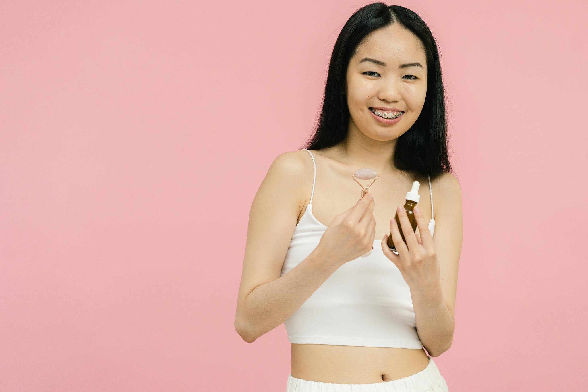 Woman smiling with facial roller and serum bottle against a pink backdrop, promoting skincare.